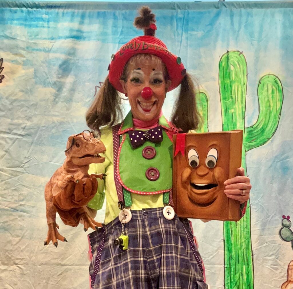 Lovely Buttons the Clown holding a T-rex puppet and Book with a fun face on it.