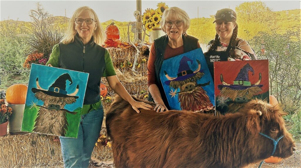 3 women holding a picture of a cow while petting a real cow.